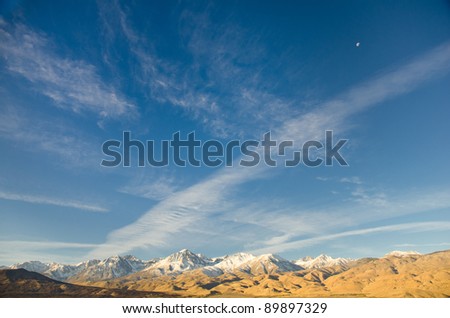 The Eastern Sierra mountain range showing big sky, clouds and the moon in daylight.