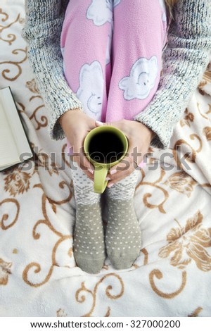 Soft cozy photo of slim tan woman in warm sweater on the bed with cup of tea in hands, top view point.Composition with warm plaid, book, cup of hot drink and female legs