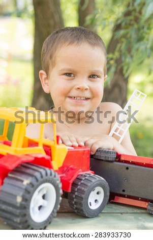 a little boy, two years old, smiling, summer, naked to the waist, sitting at a table playing with a red fire engine and a red tractor