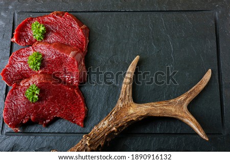 Raw steak meat from roe deer on the bridlic  chopping board. Roe deer antler as a decoration. Copy space for text.