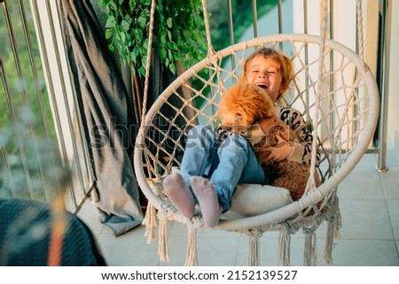 happy little girl, child hugging with a smile her pet, poodle dog at home on the balcony in spring, summer in a cotton-fringed hammock chair at sunset. The animal is like a member of the family. Imagine de stoc © 