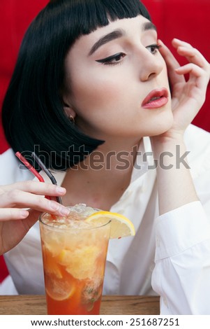 Beautiful cute woman in a black wig and the white shirt. Closeup portrait of a young cute girl over red background. Perfect bright makeup. Red lips. Pulp Fiction. With cocktail.