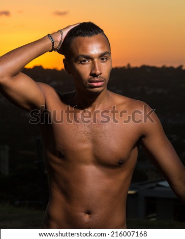 Handsome dark skin man standing torso naked in front of a cityscape at sunset