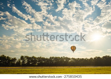 Hot air balloon flying over the road and trees in a blue sky with sun and white clouds
