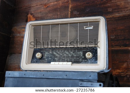 Vintage radio standing on a steel box with wooden wall in background.