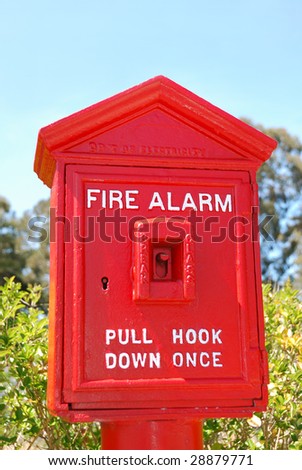 Fire alarm red box shown outside with green bushes and blue sky background.