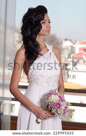 Indoor portrait of adorable bride with black long hair in front of window. Indoor portrait of adorable bride with black long hair in front of window. White long dress, wedding bouquet.