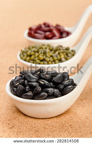 Different varieties of beans in ceramic spoons on a cork board: red kidney beans, mung beans, black beans