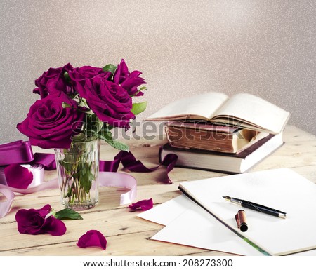 Notepad with fountain pen, red roses in a glass and old books on wooden desk in a vintage style