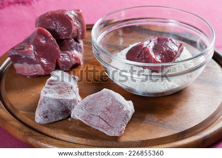 Stew meat, cover in flour meat. pieces of meat all around, flour a glass bowl. wood and a pink tablecloth in background