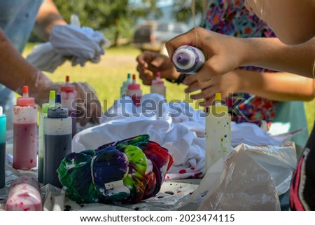 A close up of a group of people making tie dye tee shirts outside on a table- A fun day outside making tie dye shirts- Colorful bottles of dye on a table outside- A group of kids doing artwork