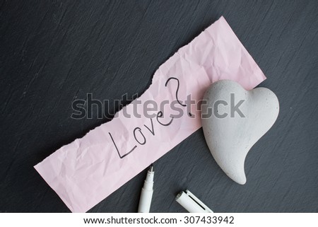 Hand-written text Love? on a piece of pink paper, one heart of stone and a pencil