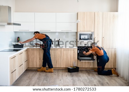 Two handymen, workers in uniform fixing, installing furniture and equipment in the kitchen, using screwdriver indoors. Furniture repair and assembly concept. Horizontal shot