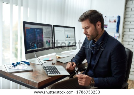 Preparing financial report. Young focused businessman or financial analyst sitting at his workplace in the office and working on laptop. Analyzing statistical data. Focus on man. Business, finance