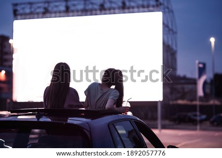 Rear view of two female friends sitting in the car while watching a movie in an open air cinema with a big white screen. Entertainment concept. Focus on people. Horizontal shot