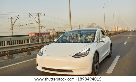 Front view of sedan electric car with driver and passenger moving along aspalted city road in sunlights, widescreen. Luxury and success concept