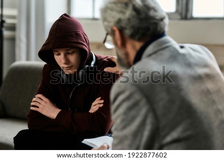 Psychotherapist supporting depressed teenage boy in hood sitting with crossed hands on couch during appointment in office, selective focus. Psychotherapy concept Photo stock © 