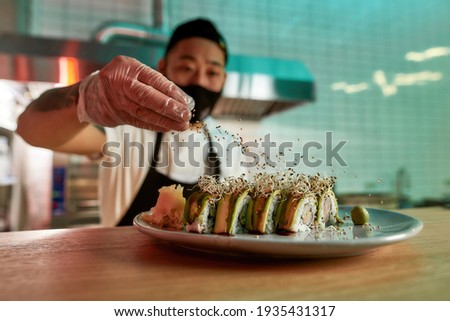 Middle aged asian man preparing sushi in restaurant. Masked chef standing in the kitchen and sprinkles sesame seeds on sushi on the table. Healthy food nibble concept in restaurant