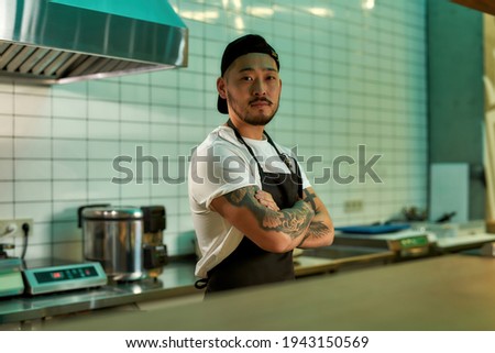 Man standing in the kitchen and looking at the camera. Young asian chef ready to cooking delicious food. Cook with folded hands posing in the cuisine. Restaurant work concept