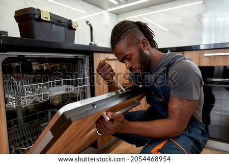 Process of dishwashing machine installation. Young African service man setting up dishwasher in new stylish kitchen. Diverce handyman in workwear, close portrait.