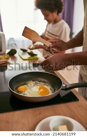 Black family cooking breakfast at home kitchen. Focus on foreground of man frying eggs on pan with background of blurred son smearing kiwi on bread piece. Relationship. Fatherhood. Domestic lifestyle Foto stock © 