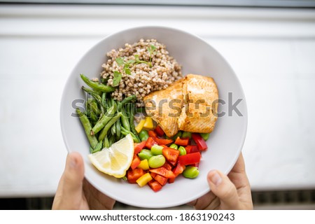 Hands holding salmon and buckwheat dish with green beans, broad beans, and tomato slices. Nutritious dish with vegetables and fish from above. Healthy balanced diet