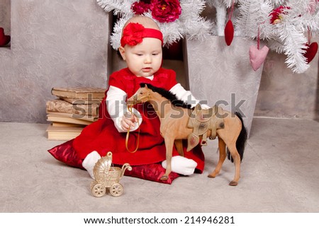 Little girl in red dress near Christmas tree with a toy horse. Baby 5 months. Christmas, New Year