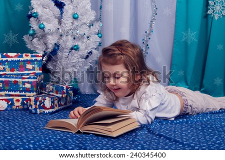 Little girl fun preschool near the Christmas tree reading a book. Blue and white interior. New Year, Christmas