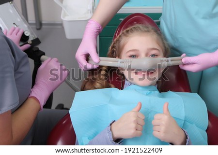 A little girl is comfortable to treat her teeth under superficial sedation. The girl smiles and holds two thumbs up. Milk teeth treatment. Treatment of children's teeth with nitrous oxide. 
