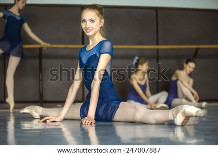 Ballerina sitting on the floor in the splits in a dance class dancers practicing on the background. Dancer is smiling.