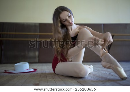 Young ballerina sitting on the floor dance classroom gently tying pointe. Beside her on the floor is a long-skirted hat, costume attribute.