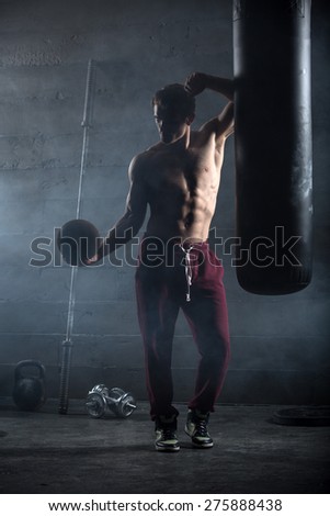 Young athlete with the ball in one hand stands near the punching bag. Snapshot in dark colors.