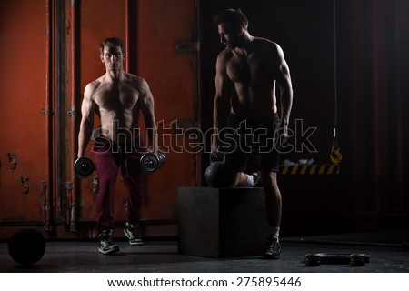 Two beautiful athlete train. One does kettlebell swings, squats with dumbbells other. The picture in the studio in a dark tone.