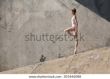 Slim dancer stands in a ballet pose and looking down on a gray urban concrete background. Outdoors shooting with sun light.