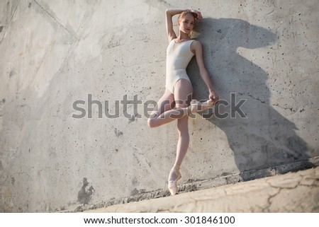 Slim dancer stands in a ballet pose and looking down on a gray urban concrete background. Outdoors shooting with sun light.