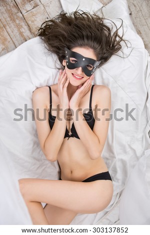 Playful beautiful brunette with curly hair in a black leather mask lying on her back on the bed and looking up at the camera with a smile.