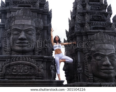 Young woman in white dress doing yoga asana at old temple surrounded by face of Bayon