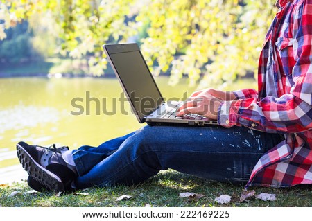 Young man with laptop near lake in city park outdoor. Caucasian man sitting on green grass and working on his laptop