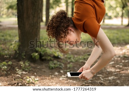 Young attractive girl hanging upside down in the city park and holding white tablet pc in hands. Smiling curly woman outside having fun and talk with friends while using 5G internet. 商業照片 © 