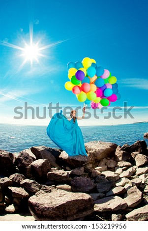 Luxury fashion woman with balloons in hand on the beach against the sky and the sun in long dress
