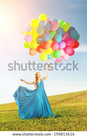 Luxury fashion stylish woman with balloons in hand on the field against the sky and  the sun in long dress