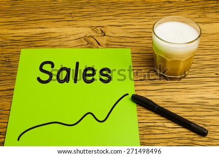 Sales written on green paper with graph. Marker and coffee on desk.