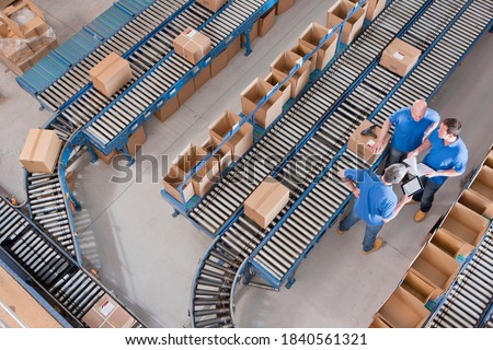 Top view of workers with papers and a digital tablet having a discussion among boxes laid on conveyor belts at a distribution warehouse.