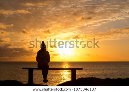 Wide angle shot of the Silhouette of a woman sitting on a bench looking at the tranquil sunset over the ocean