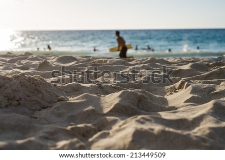 sand close-up at beach near blue ocean where people are swimming and surfer is running to catch waves