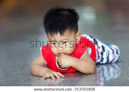 The baby Asian male wearing a red shirt. Crawling, playing on a cement floor inside the house.