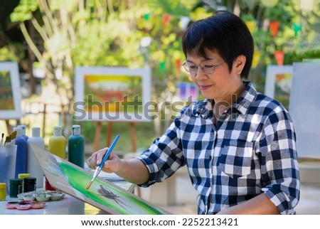 talented female artist sitting and holding paint brush painting watercolor on painting pad, middle aged asian woman having inspiration while working on a picture 
