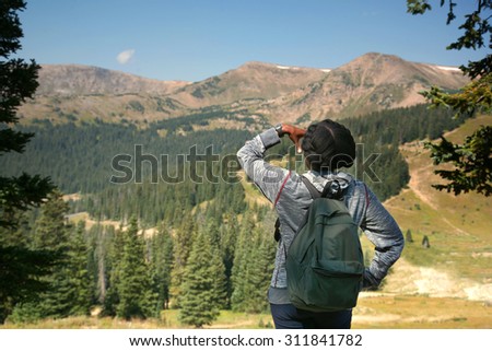 Young black female tourist views mid-day landscape of mountains with evergreens and deep blue sky