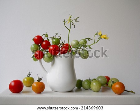 Organic green and red tomatoes in a white pitcher still life on a light background. With tomato flowers.