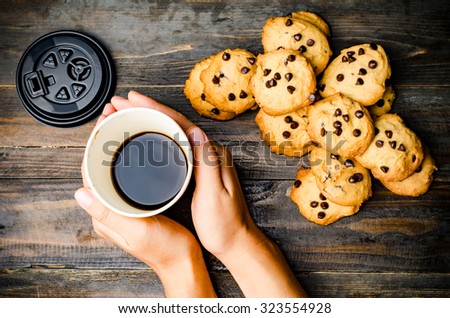The cup of hot coffee is holding by hand and chocolate chip cookies on wooden background